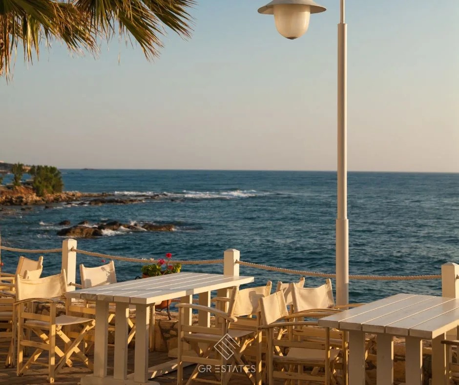 A seafront café terrace at golden hour, with white wooden tables and director's chairs behind a rope railing, a palm frond, a street lamp, red geraniums and a calm blue sea breaking on rocks